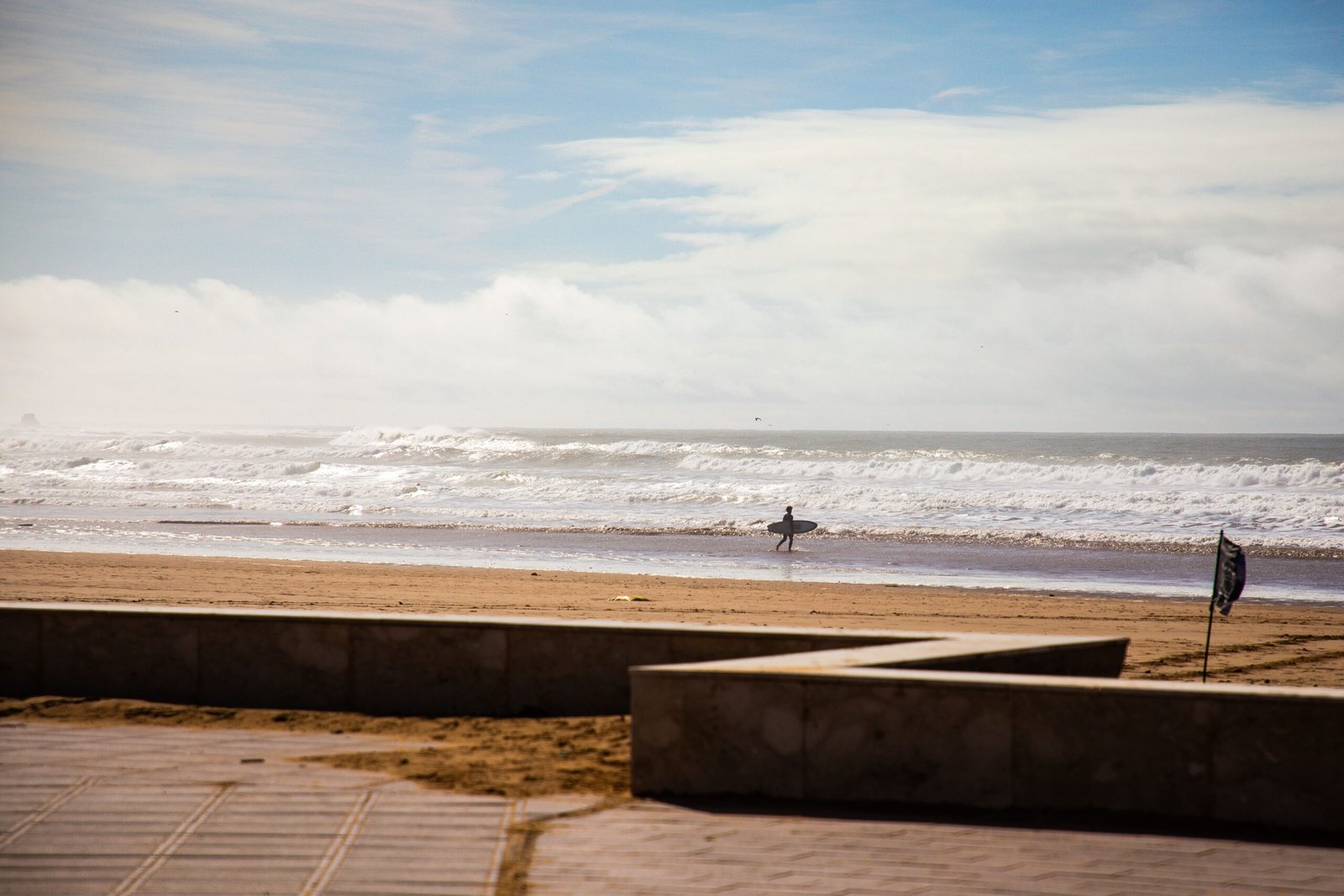 A lone surfer walks along a sunny Moroccan beach with waves and blue sky in the background.