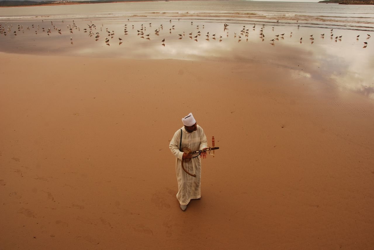 A man in traditional Moroccan clothing plays an instrument on a serene beach.